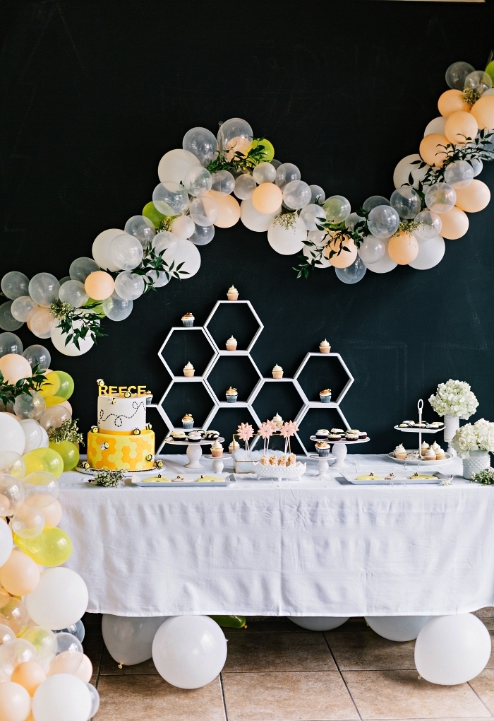 theme anniversaire abeilles en noir, jaune et blanc pour fêter le premier an de votre bébé, une déco de table d'anniversaire avec arche de ballons et de feuilles artificielle, buffet d'anniversaire avec bonbonnières et présentoir à cupcakes en forme de nid d'abeille décoré d'une arche de ballons sur un fond noir