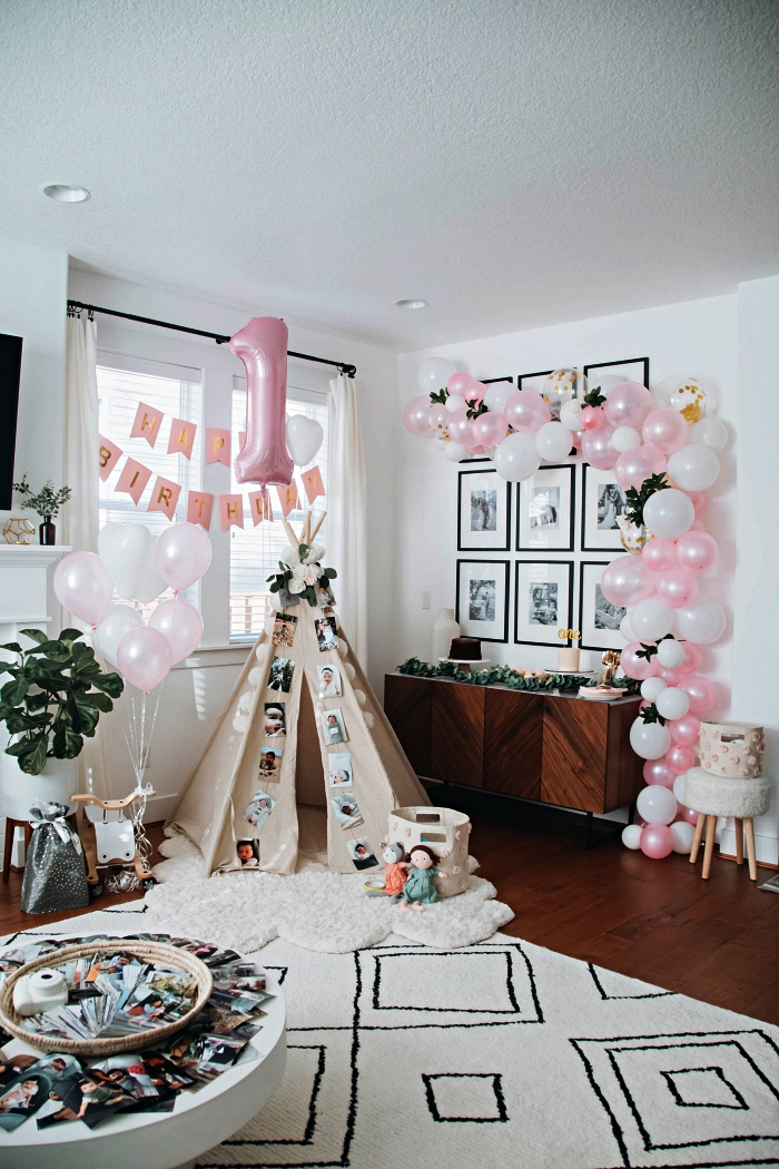 déco d'anniversaire enfant bohème chic avec tipi dans le salon, une arche de ballons en rose et blanc, ballon chiffre aluminium et une banderole joyeux anniversaire 1 an