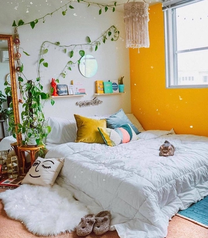 ambiance cosy dans chambre boheme chic, mur d accent peinture jaune, lit bas décoré de coussins colorés, plante verte grimpante d interieur, tapis peau de mouton, miroir rectangulaire vintage