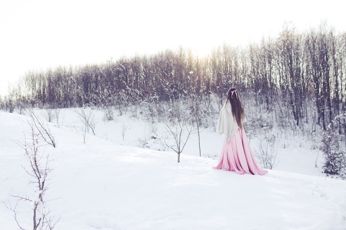 idee de femme qui marche à travers un champ enneigé avec des arbustes et arbres bas sans feuillage, image feerique femme robe rose et cape blanche