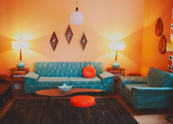 murs repeints de couleur orange interieur, canapé et fauteuil bleu, tapis marron foncé, coussin et tabouret orange, eclairage interieur original