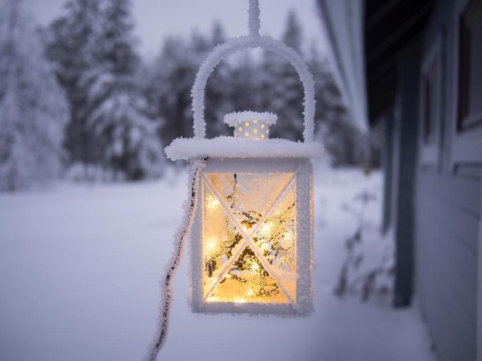 lanterne blanche remplei de guirlandes lumineuses pour decorer l exterieur d une maison grise, paysage de neige dans cadre forestier