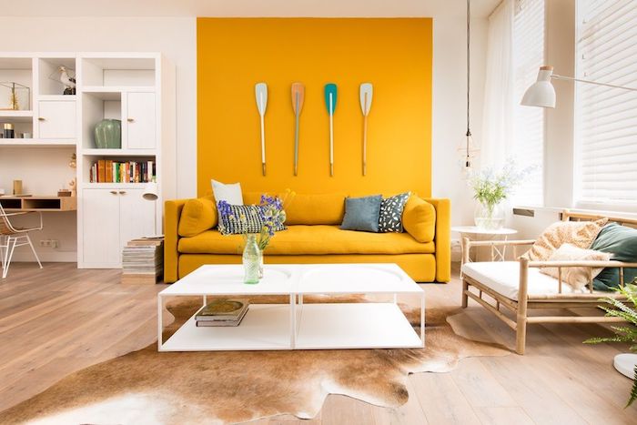 pan de mur couleur jaune vive et canapé jaune dans un salon blanc, deco murale originale en avirons, parquet bois clair et chaise de bois, tapis peau d animal