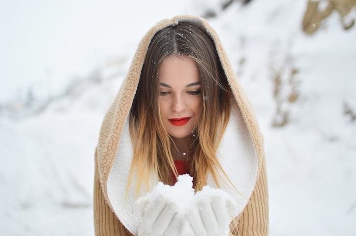 image hiver de femme avec manteau au capuchon qui tient dans ces mains de la neige, fond pittoresque enneigé
