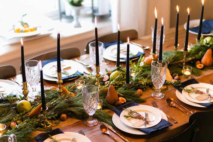 chemin de table noel en branches de pin, noix, poires et autres fruits, chandelles en ros avec bougies noires, serviettes bleu marine sur table bois blond