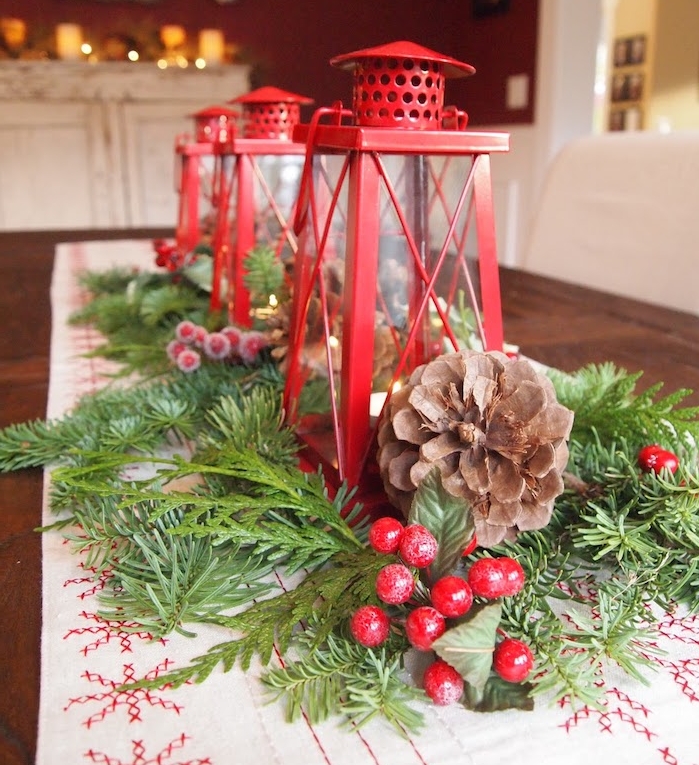 centre de table de noel de couleur blanche à décorations rouges avec feuillages de pin, lanternes rouges, houx et pommes de pin