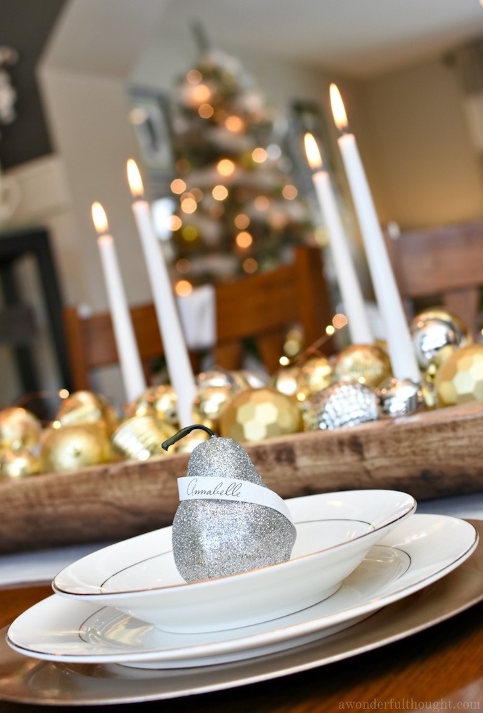 marque place en poire pailletée dans assiette blanches, centre de table en bois avec déco de boules de noel or et argent et bougies blanches