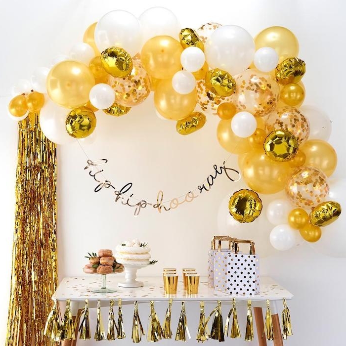 arche de ballons en or et blanc sur mur fond blanc, deco guirlande de pompons à franges dorées, candy bar de fete avec beignets et gateaux