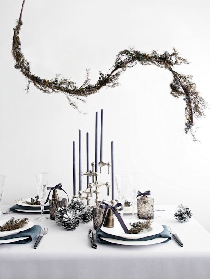 decoration scandinave de nouvel an, branche decorative au dessus d une table à nappe blanche, bougeoir et pommes de pin argentées, motifs deco vegetation