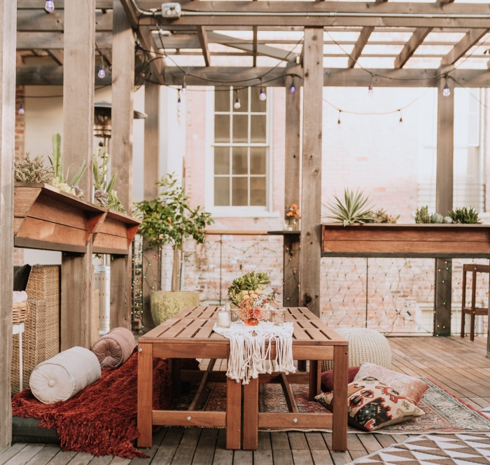 anniversaire-style-bohème-chic-table-bois-décoration-party-extérieure-nappe-macramé-coussins-par-terre-plantes-vertes-guirlande-lumineuse modèle de centre de table anniversaire avec bouquet de fleurs fraîches, idée déco naturelle et bohème sur une terrasse bois