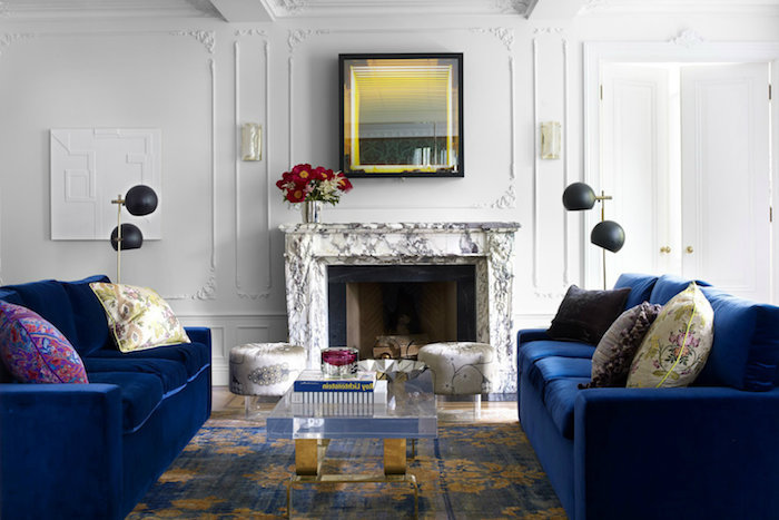 cheminée de marbre dans un salon blanc avec canapés bleu marine, table basse en verre transparente sur tapis or et bleu, lampes noires