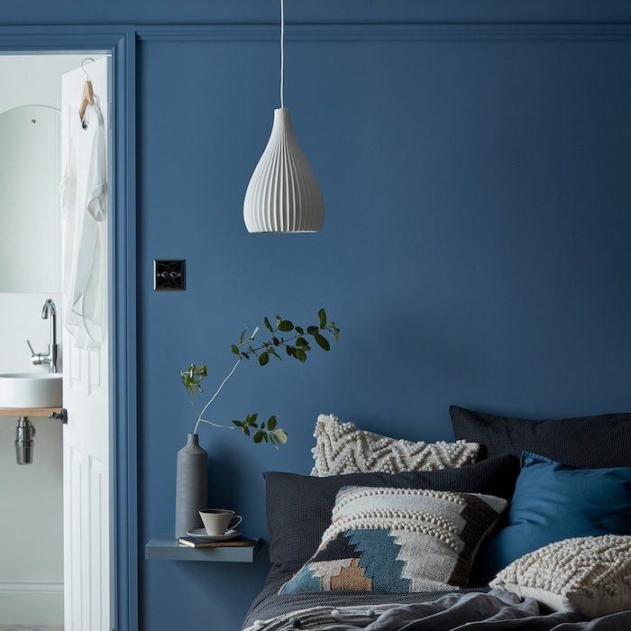 mur pantone bleu couleur interieur, coussins de tons foncés sur un lit avec linge de lit gris, suspension blanche