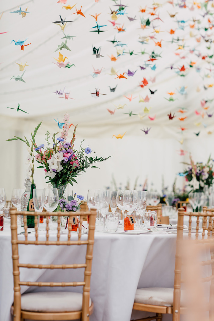origami papier coloré grues suspendues du plafond et décoration de bouquet de fleurs champetres colorées sur nappes blanches, decoration salle mariage colorée
