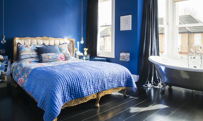 baignoire gris clair dans une chambre à coucher adulte bleu foncé, lit baroque avec linge bleu à imprimé floral, parquet noir