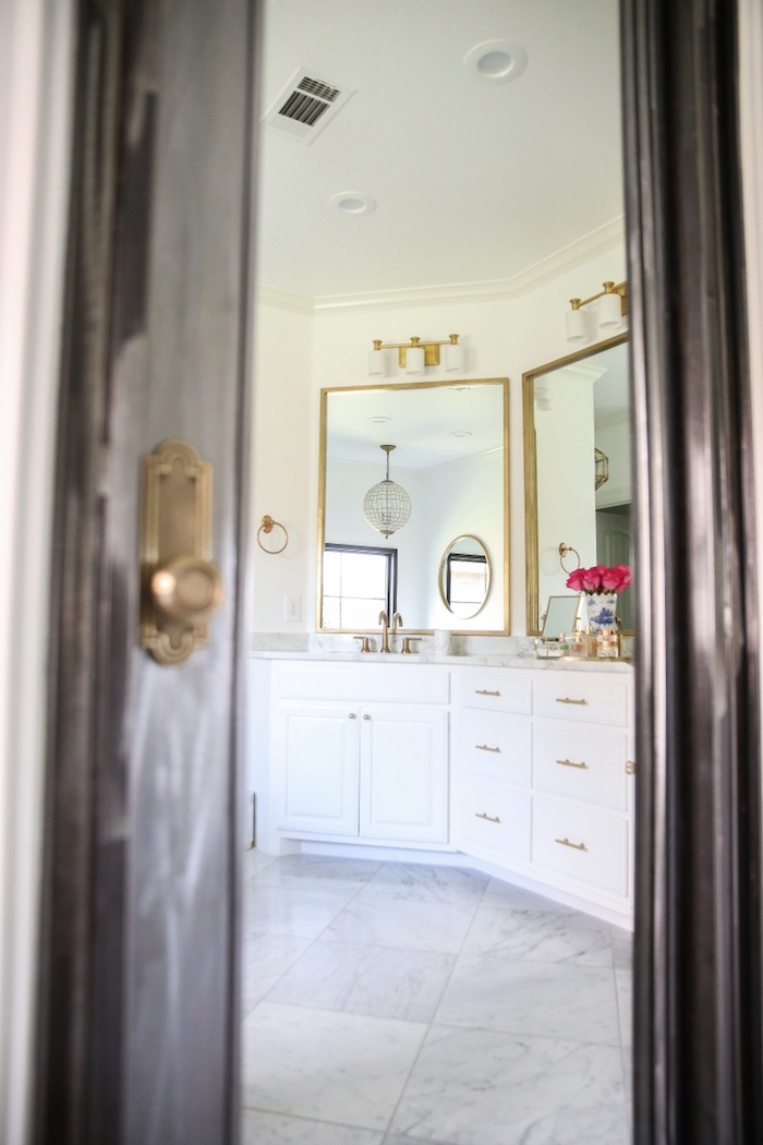 Vue de la porte ouverte à la salle de bains avec sol en marbre, peinture murale blanche, miroirs dorés, meuble lavabo blanc bien rangé, fleurs dans une vase