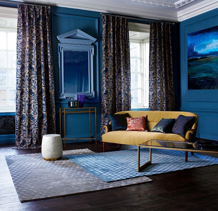 canapé jaune décoré de coussins dans un salon bleu foncé avec tapis gris et tapis bleu sur parquet bois foncé, rideaux gris et bleu