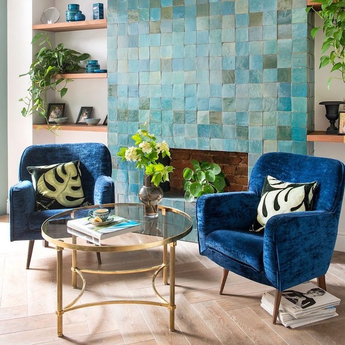 fauteuils bleu marine dans un salon avec cheminée bleu clair carrelage, étagères de bois ouvertes, accents bleu paon, table verre et laiton