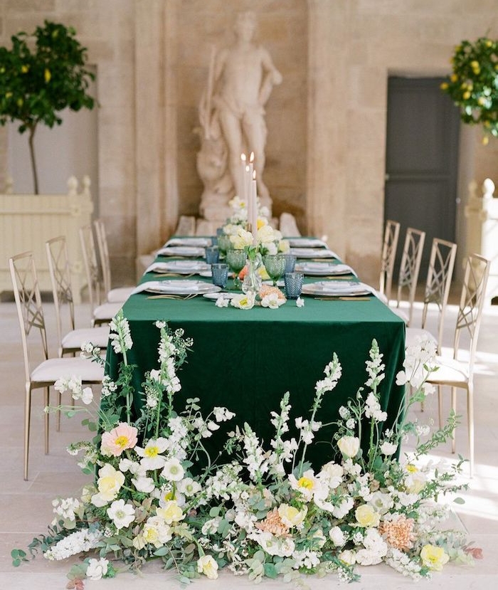 verres de couleur bleue et assiettes blanches sur nappe verte, table entourée de chaises argent et blanc, pieds de table fleuris, statuettes romaines