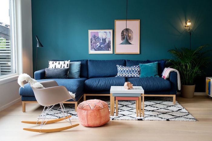 canapé en bleu classique et mur en bleu paon dans un salon scandinave cocooning avec chaise à bascule scandinave, pouf saumon, tapis noir et blanc