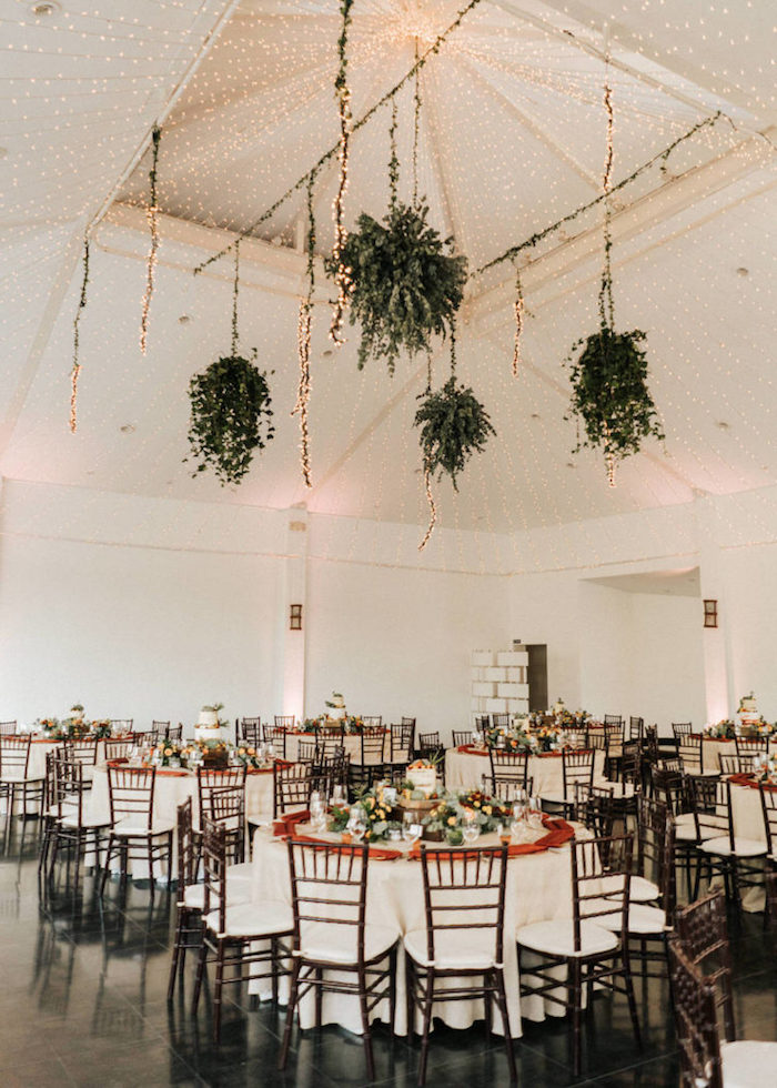 pots de plantes vertes retombantes suspendues et plafond décoré de guirlandes lumineuses au dessus de tables en nappes blanches et chaises bois et blanc sur sol carrelage noir