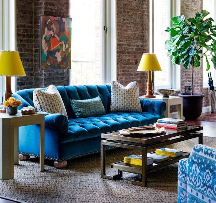 mur de briques et grandes fenetres dans salon style industriel avec canapé bleu et table bois originale sur tapis gris et blanc, plante verte en grand pot