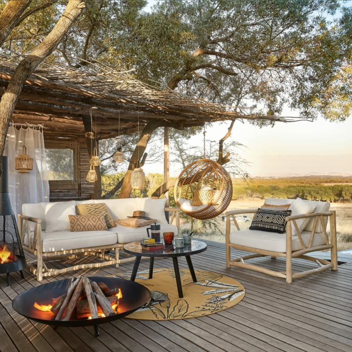 Belle vue d'une terrasse bien aménagée, canapé bois et coussins blanches, diy cheminée tapis ronde jaune, balançoire oeuf chaise
