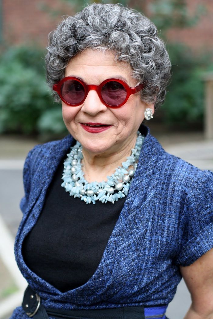 exemple de coupe de cheveux court femme 60 ans avec lunettes monture rouge cheveux gris bouclés collier bleu veste bleue cheveux volumineux
