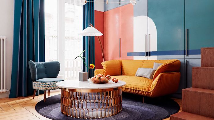 canapé jaune moutarde fauteuil bleu table basse marbre et laiton tapis rond gris rideaux bleus armoire bleu blanc et orangé parquet bois clair