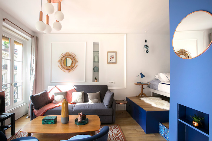 deco studio 20m2 avec canapé gris table basse bois fauteuils bleus tapis rouge et gris lit sur estrade mur d accent bleu