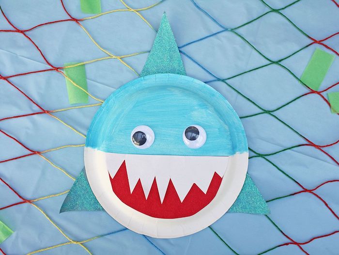 requin fabriqué en assiette de papier blanc et bleu avec bouche rouge et des avirons bleu pailletés des yeux mobiles acticité pour les enfants