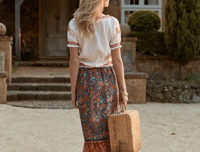tenue aesthetic style boho chic une femme blonde devant une maison rustique vetue en jupe maxi florale portante une valise