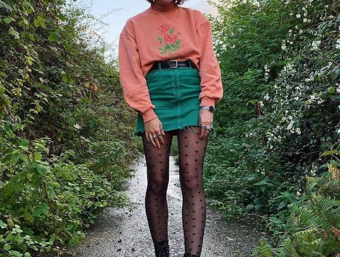 tenue stylée femme des années 90s soft grunge aec une jupe courte en jean vert pull rose et bottes noires avec un collant a pois dans la nature
