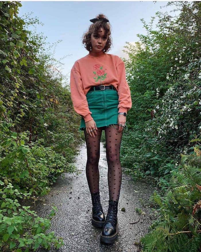 tenue stylée femme des années 90s soft grunge aec une jupe courte en jean vert pull rose et bottes noires avec un collant a pois dans la nature