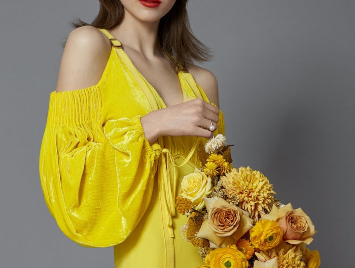 une femme qui tient un bouquet de fleurs oranges vetue en robe jaune au fond gris