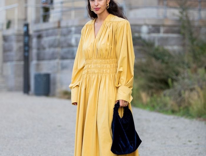 une femme vetue en robe longue jaune avec un sac a main noit devant un edifice culturel