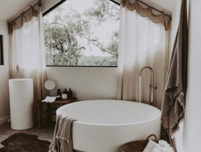 baignoire blanche dans salle de bain blanche et grise avec tapis marron foncé panier rempli de serviettes blanches sdb sous comble