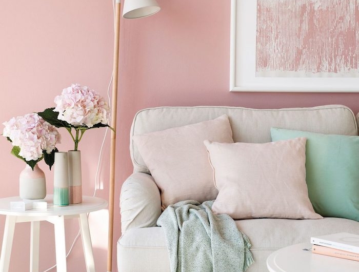 un salon aux murs roses derrière un canapé beige décoré des coussins vert menthe et une peinture rose poudré qui pend