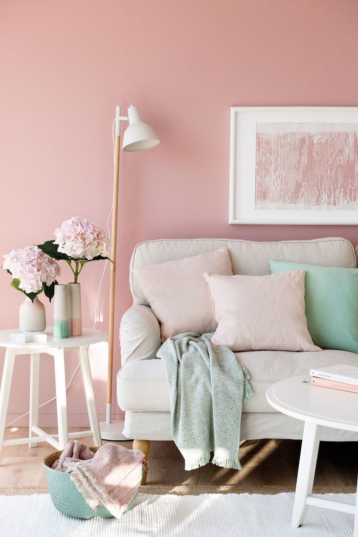 un salon aux murs roses derrière un canapé beige décoré des coussins vert menthe et une peinture rose poudré qui pend