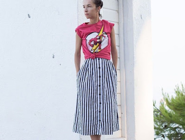 une femme qui porte une jupe longue a rayes et un t shirt rose avec un imprimé devant un batiment blanc