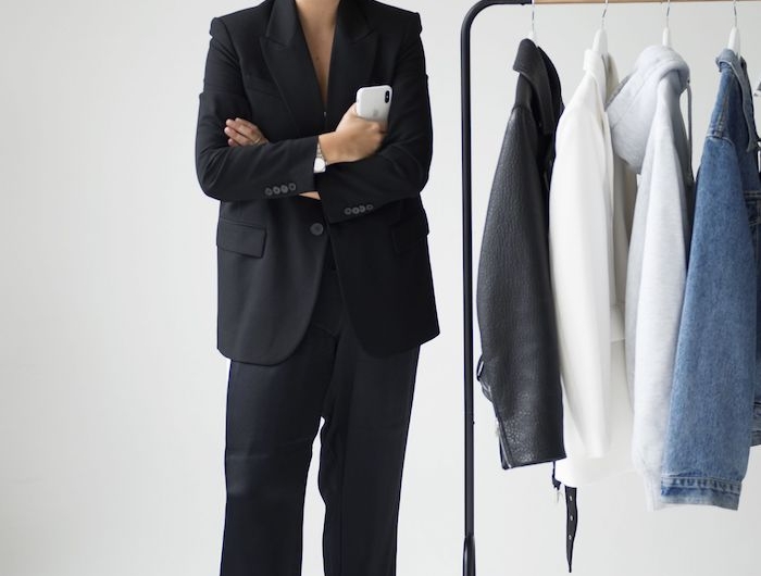 une femme vetue en tailleur noir classique e coté d un porte manteau ou garde robe minismaliste