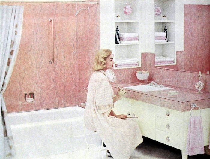 une salle de bain des années 50 avec un carelage couleur rose pastel et une femme assise devant le miroir
