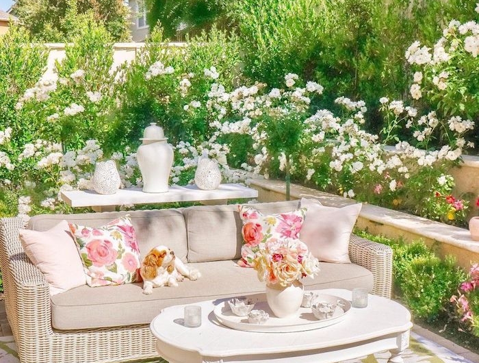 aménager un jardin planté des fleurs avec des canapés en rotin et des coussins beiges avec un petit chien au dessus