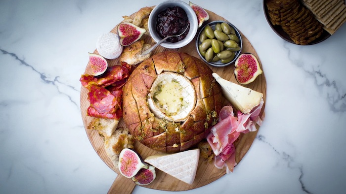 idéee de recette avec camembert dans un pain entourée de charcuterie olives et fruits sur une surface en marbre copy