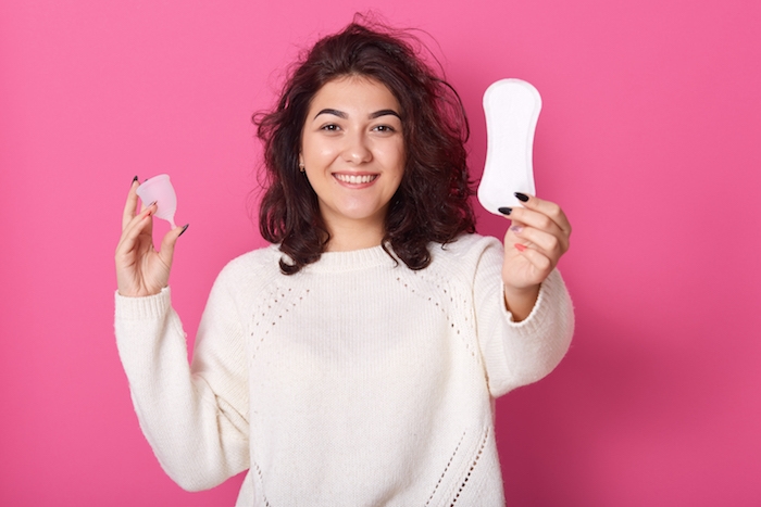 portrait of cheerful positive curly haired woman standing isolated over pink background in studio, holding hygiene pad and menstrual cup in both hands, looking directly at camera. menstruation concept