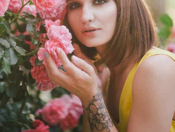 une femme en top jaune avec avec tatouage fleur poignet devant un rosier