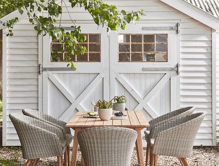une table en bois devant le garage et des chaises en rotin peints en gris