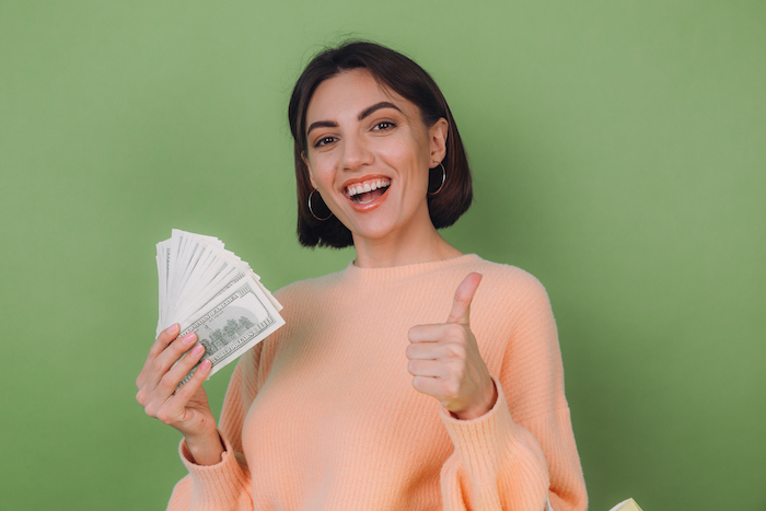 young woman in casual peach sweater isolated on green olive background holding fan of 100 dollar bills money and shopping bags copy space thumb up