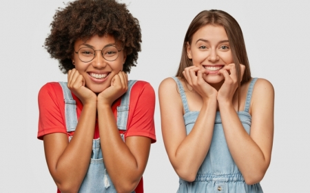 people and diversity concept. cheerful mixed race women hold chins, have toothy smiles, dressed in denim overalls, stand next to each other, isolated over white studio wall, express positiveness