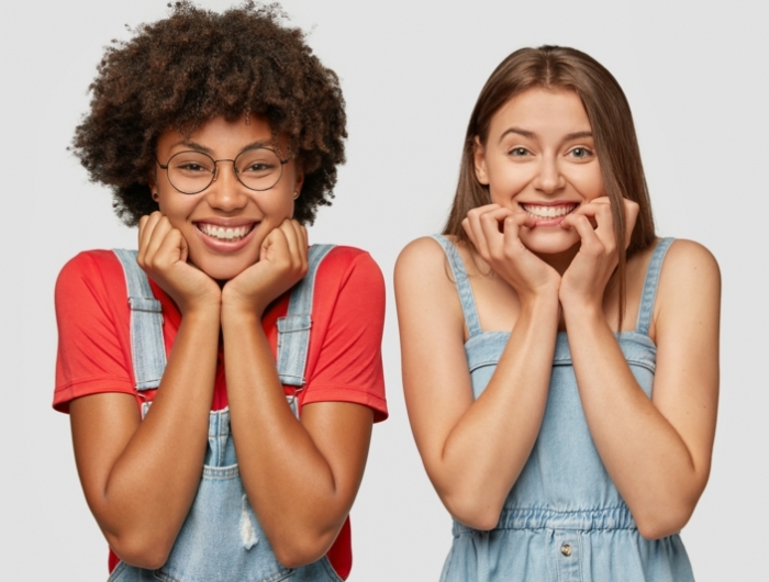 people and diversity concept. cheerful mixed race women hold chins, have toothy smiles, dressed in denim overalls, stand next to each other, isolated over white studio wall, express positiveness