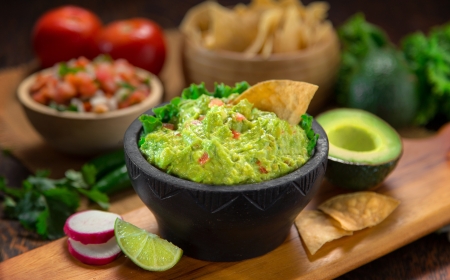 a delicious bowl of guacamole next to fresh ingredients on a table with tortilla chips and salsa